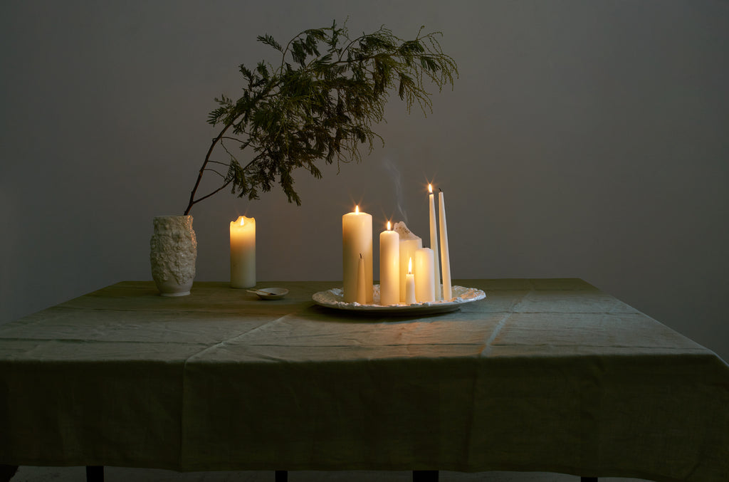 Francis Palmer relief flower vase with branches of evergreen arching over relief flower platter holding an array of white lit candle shapes and sizes on table covered with linen moss green tablecloth.