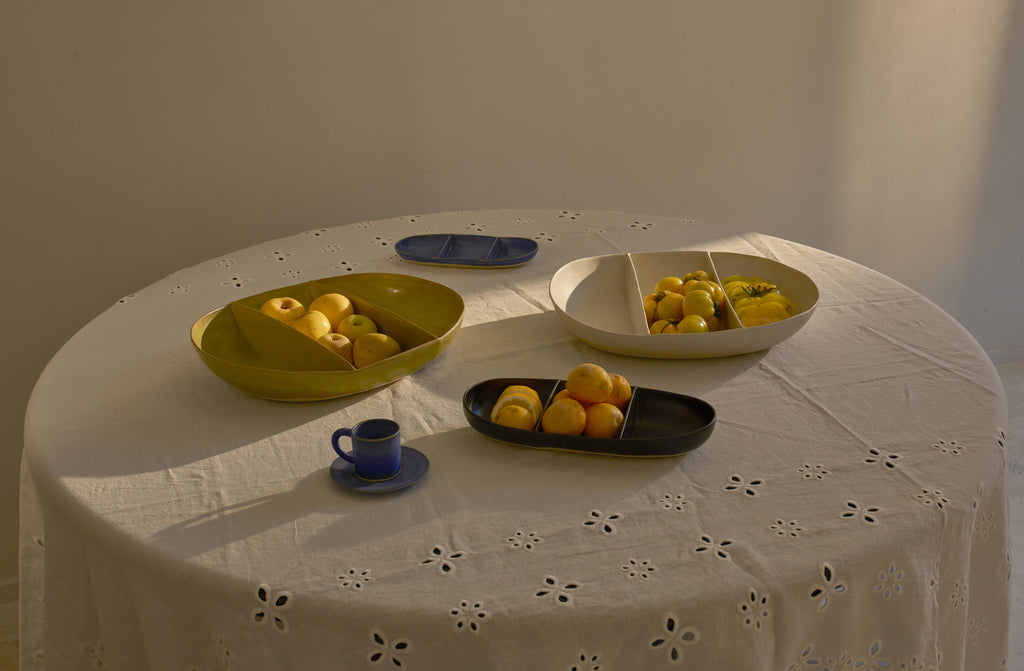 Large and small yellow-green indigo and white-beige Christiane Perrochon divided dishes with cheery yellow tomatoes and citrus on a round table covered with white Italian sangallo tablecloth.