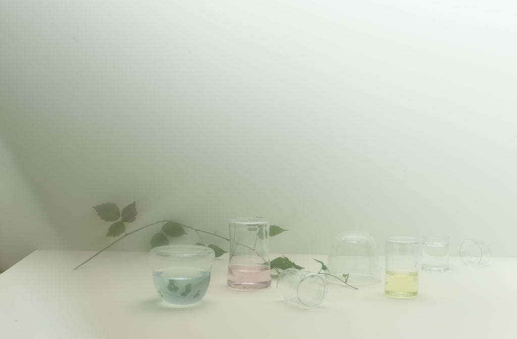 Dreamy image of Lobmeyr lidded carafe and small and round lidded boxes filled with pale pastel liquids and set on a table with a single tapering vine.