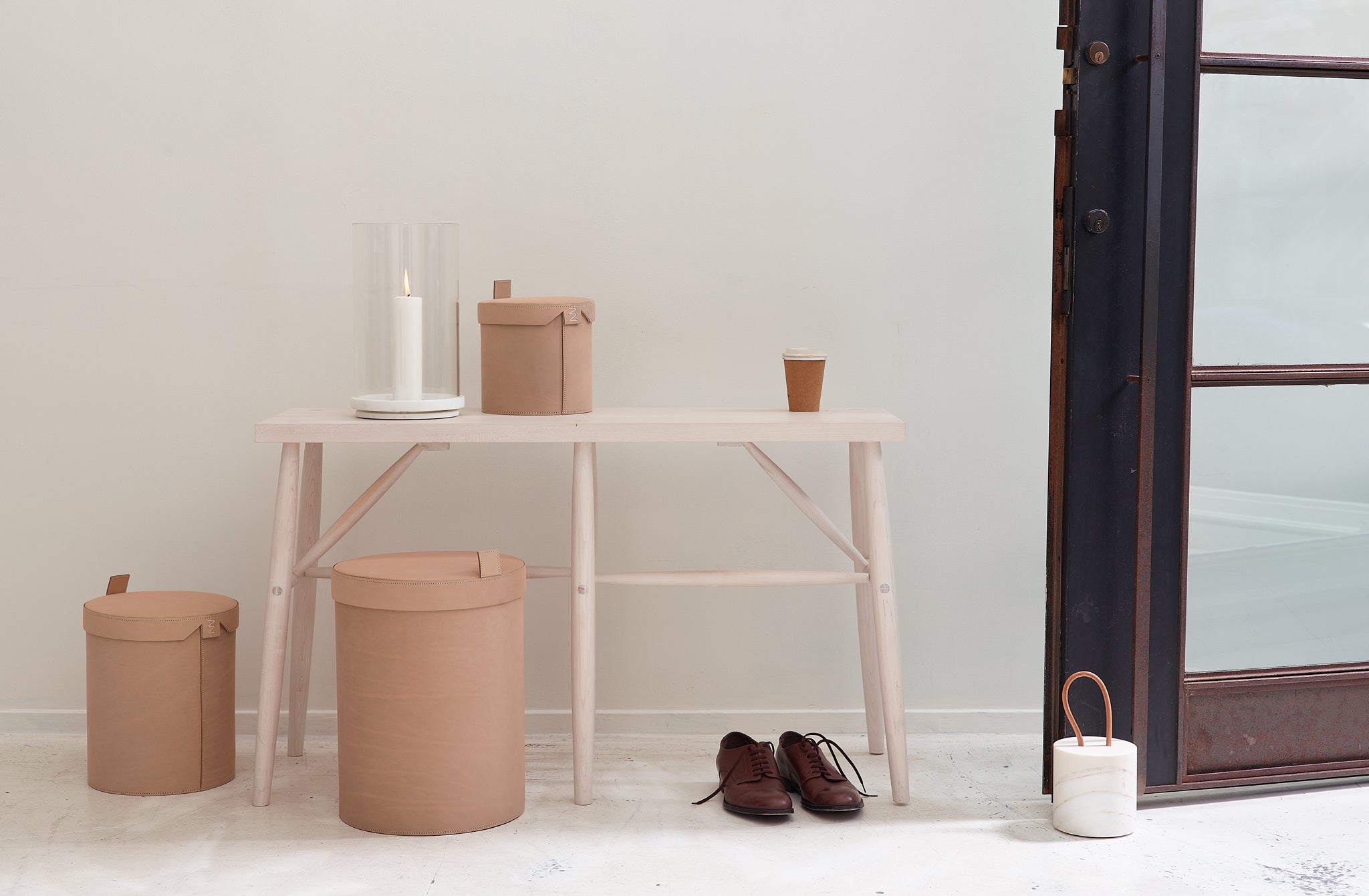 Sawkille bleached maple bench at entrance of a white room, open patinaed steel door held in place by Michaël Verheyden white marble doorstop, cement colored leather storage bins to one side with a pair of brown oxfords set under the bench.