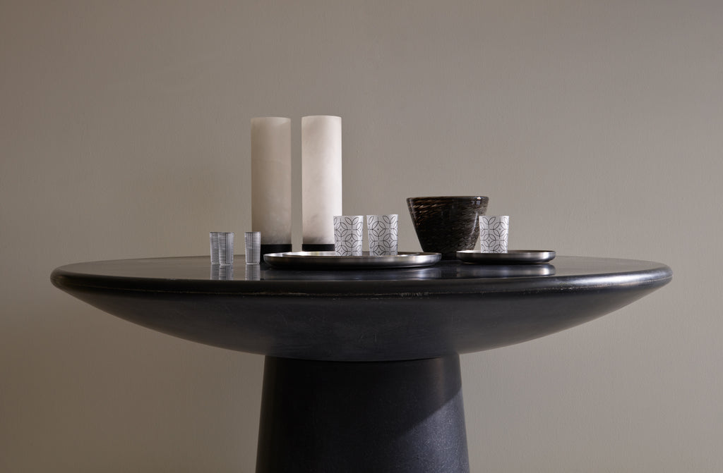 Faye Toogood charcoal Roly Poly round dining table set with a pair of Michaël Verheyden alabaster taper hurricanes and a round stainless steel serving tray set with Lobmyer hand painted tumblers with an antique grey and black murano bowl behind.