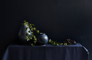 christiane perrochon indigo large and boule vases with trailing vine with dark berries set atop a navy linen tablecloth with a dark background