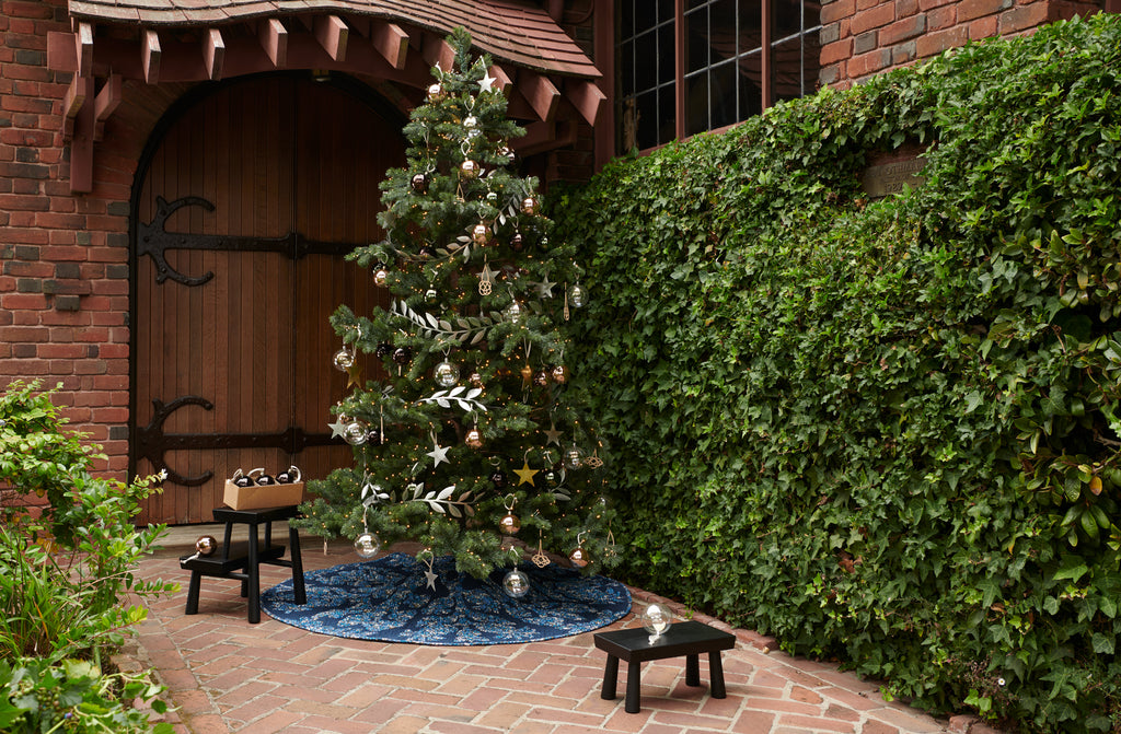 large decorated christmas tree with quilted liberty print tree skirt shown outside an old building with a massive door with iron hinges being decorated and ornaments set on black wood stepstools