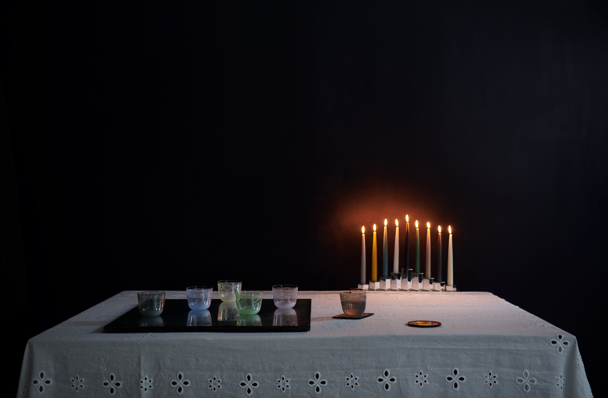 lobmeyr viennese lace engraved tumblers in various colors set on a mirrored surface with a lit candlelobra shown behind with multicolored tapered candles