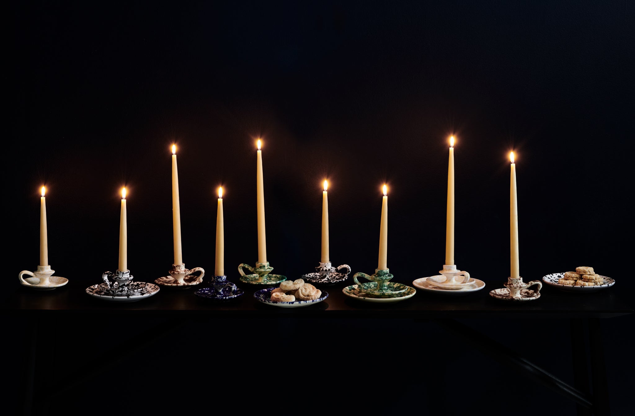 Multicolored splatterware candleholders with lit taper candles enmasse against a dark background