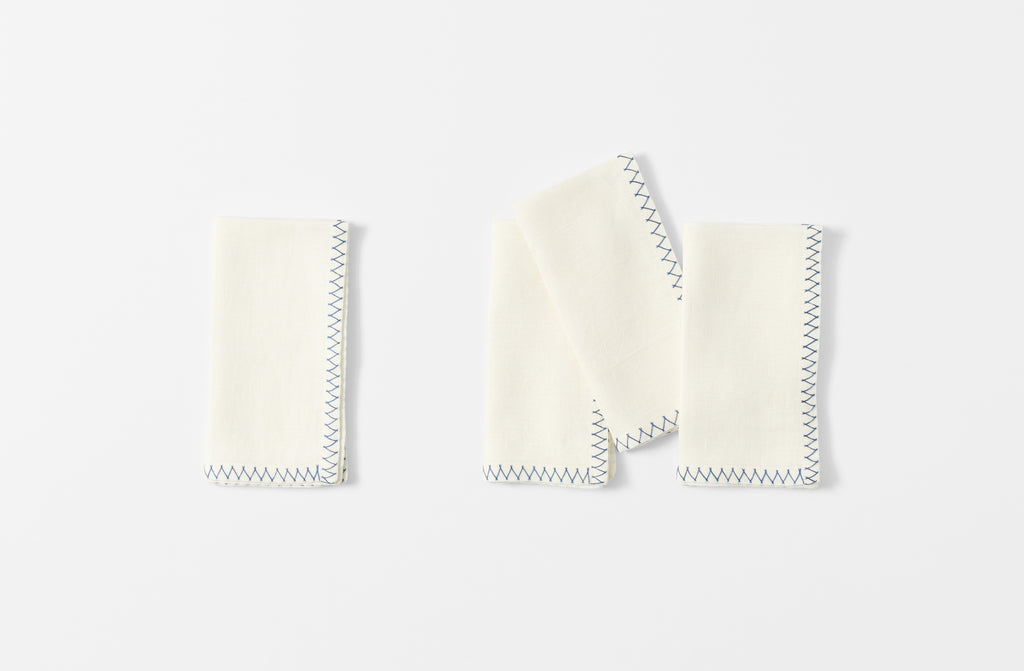 four white malaika napkins with a zig zag blue hand embroidered edge shown folded