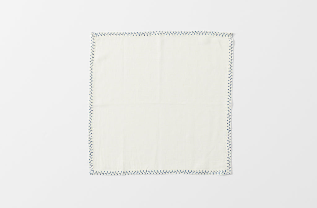 one white malaika napkin with a zig zag blue hand embroidered edge shown unfolded from above