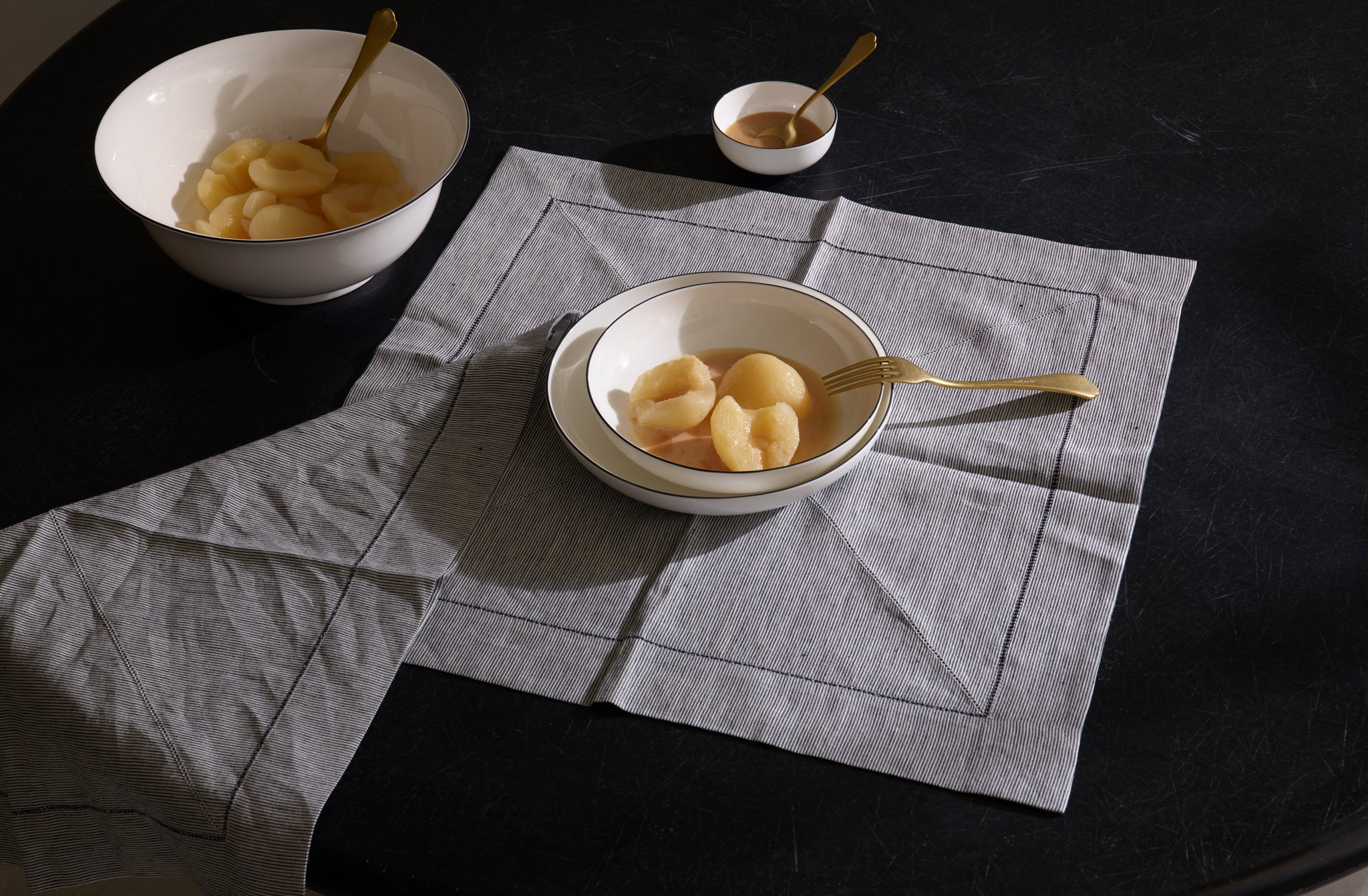 Blackline dinnerware and serving bowls from Richard Brendon set for a single serving of poached pears with Dolcevita gold flatware atop a Peter Speliopoulos navy ticking stripe linen napkin.