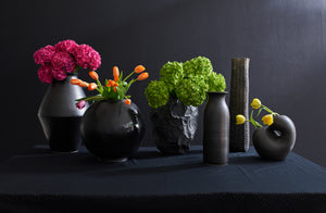 Black vessels both round and cylindrical from Faye Toogood Christiane Perrochon and Max lamb set against a black wall with neon bright flowers.