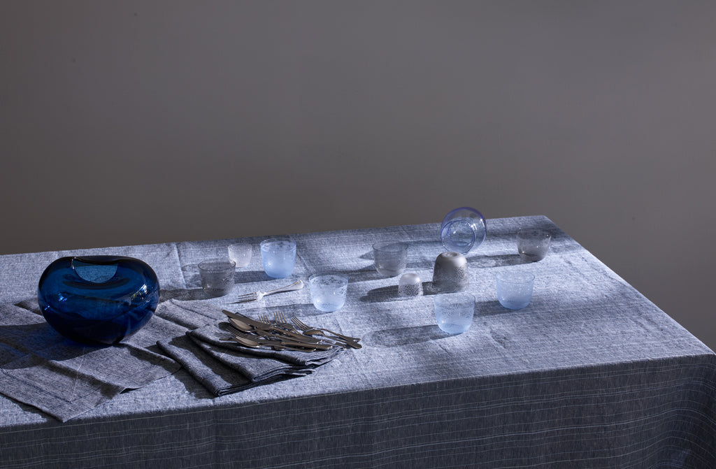 Kate Hume's night blue glass vase set tabletop with Lobmeyr lace alpha tumblers in blue and grey atop a Chiarastella Cattana Greta blue striped linen tablecloth with a grey background.