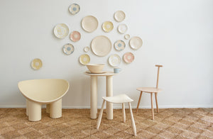 Room set with pale table, stools and chairs by Abigail Castañeda and Faye Toogood, table set with cream and blue tabletop by Brickett Davda, decorative plate wall of pastel flower plates and cream splatterware on the wall behind.