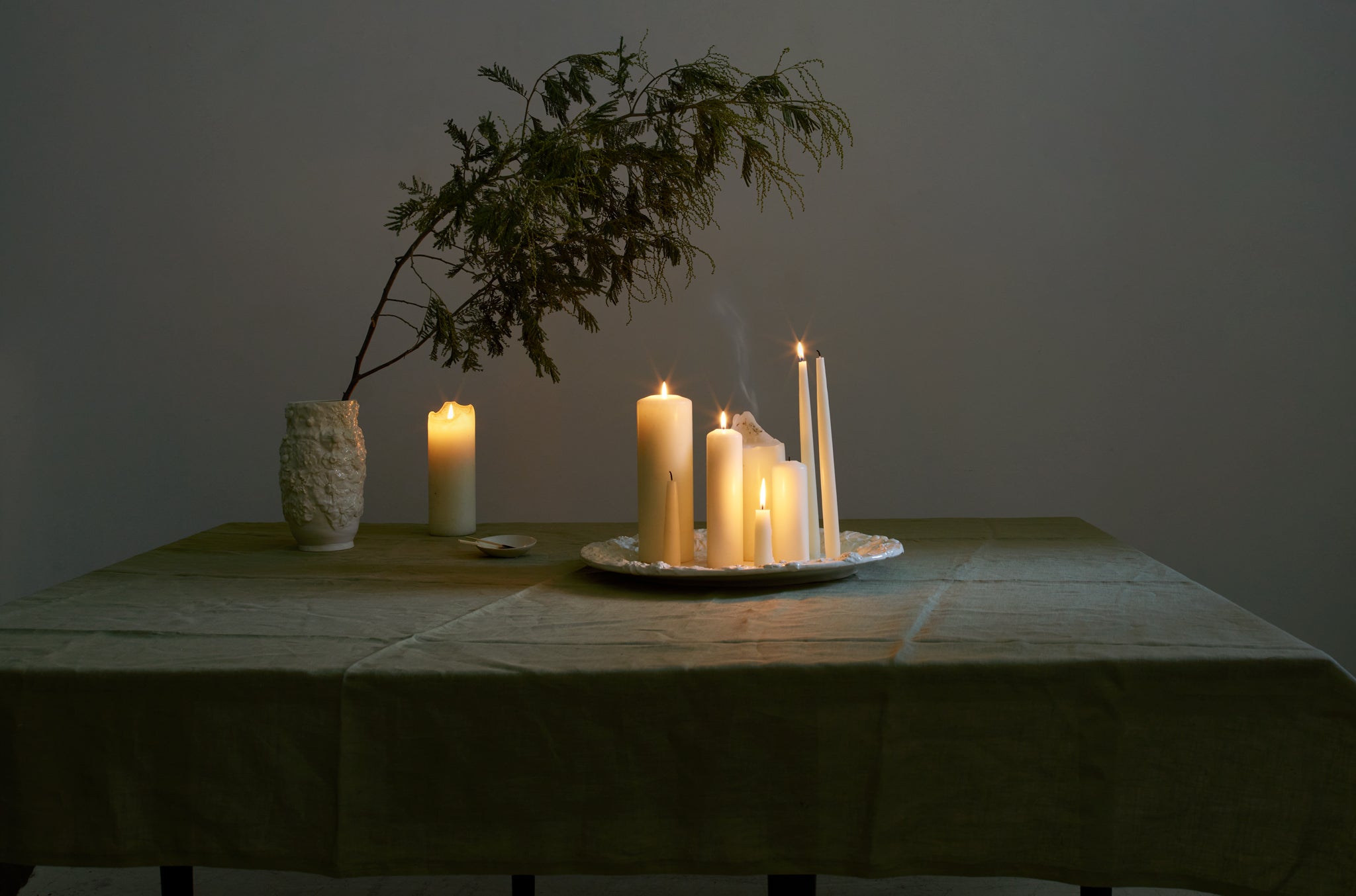 Francis Palmer relief flower vase with branches of evergreen arching over relief flower platter holding an array of white lit candle shapes and sizes on table covered with linen moss green tablecloth. 