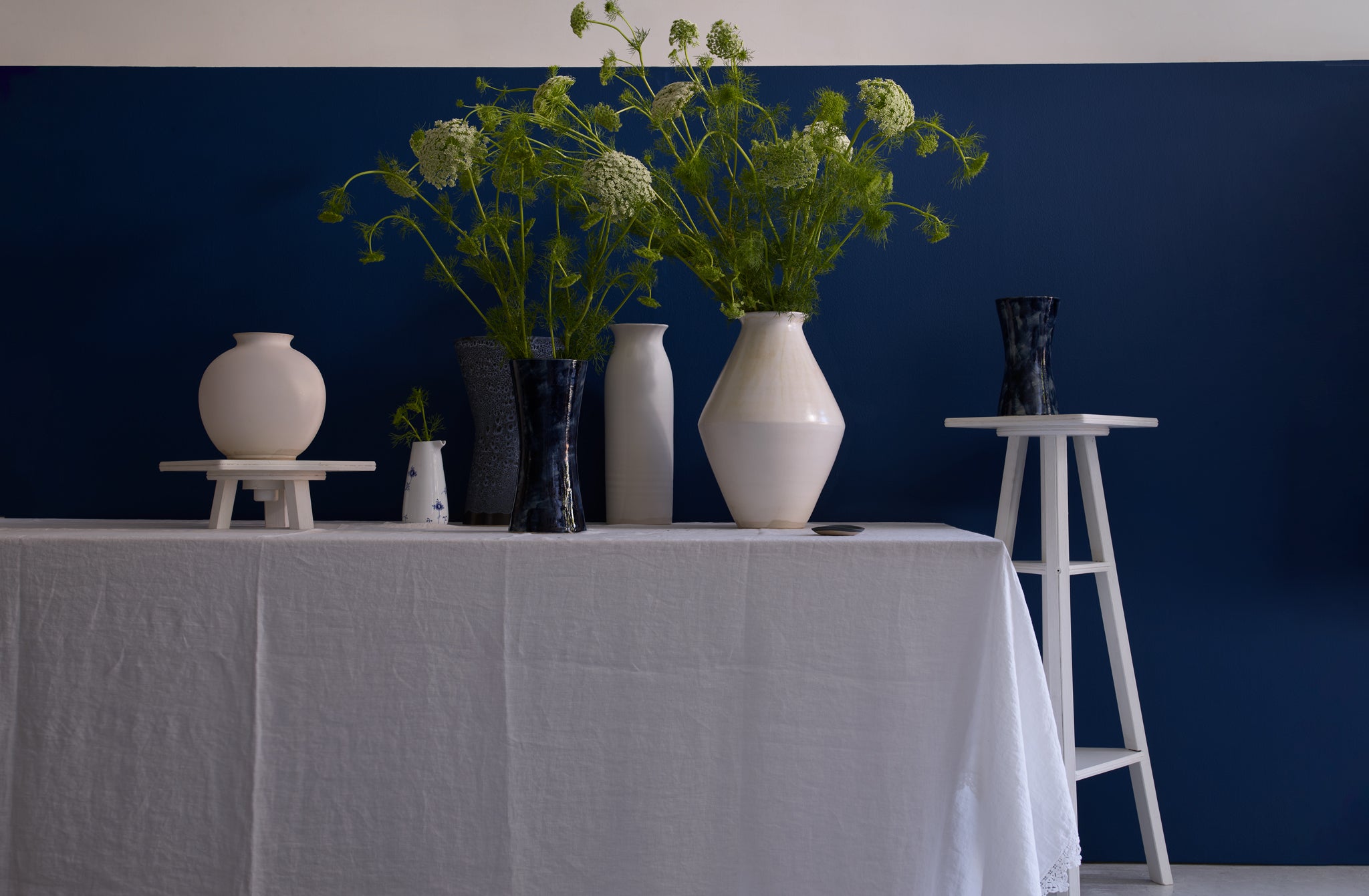 Table drapped in a white linen tablecloth and set with a tableau of Christiane Perrochon white and indigo painted vases with queen anne's lace floral against a dark blue background. 