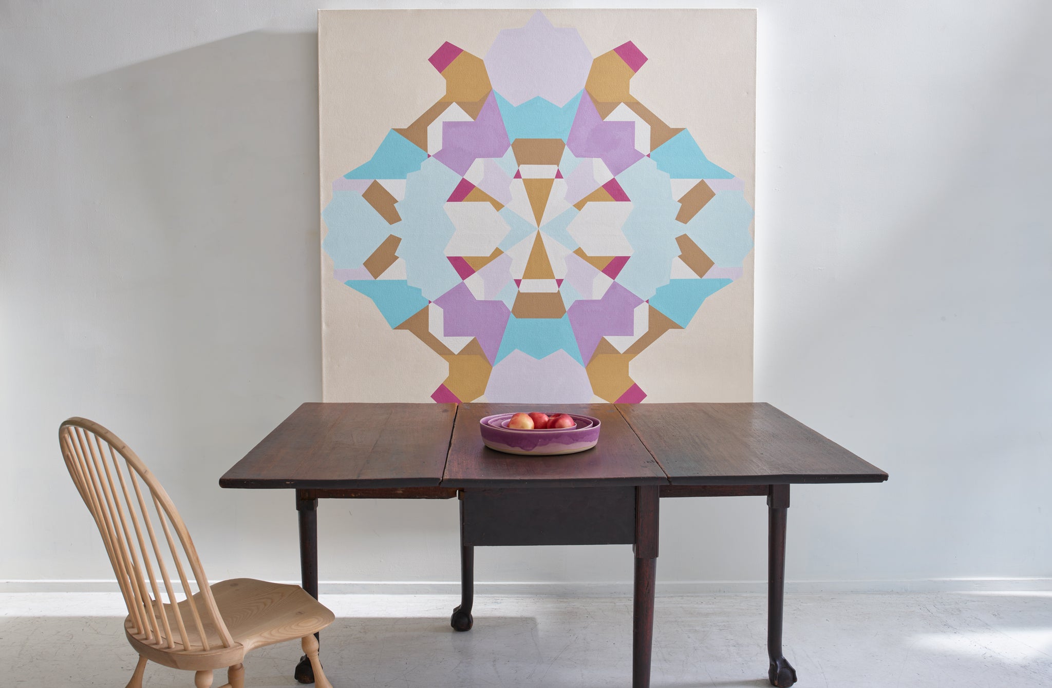 Leslie Wilkes geometric painting displayed above an antique 19th century Mallorca table set with nested Christiane Perrochon Fuschia deep dishes holding apples next to a Sawyer Made ash Windsor chair.