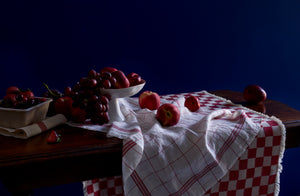 Red accented linens and red rimmed Feldspar Studio fruit bowl with a bounty of red fruit on antique table against a dark indigo blue wall.