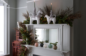 Mirrored Victorian mantle with small and large Equestre ceramic candleholders nestled in garland and Christiane Perrochon petite boules with sprigs of holiday greenery.