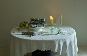 A festive scene on a Faye Toogood Roly Poly dining table covered with an off white Sicily tablecloth and a stack MARCH green marbled celebration crackers on bianco centerpiece pedestal next to a paper and ribbon packaged panettone and green milk coppa glasses.