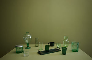 A mix of glassware for every need in shades of green from light to dark on sage green surface with a mossy green background. Default