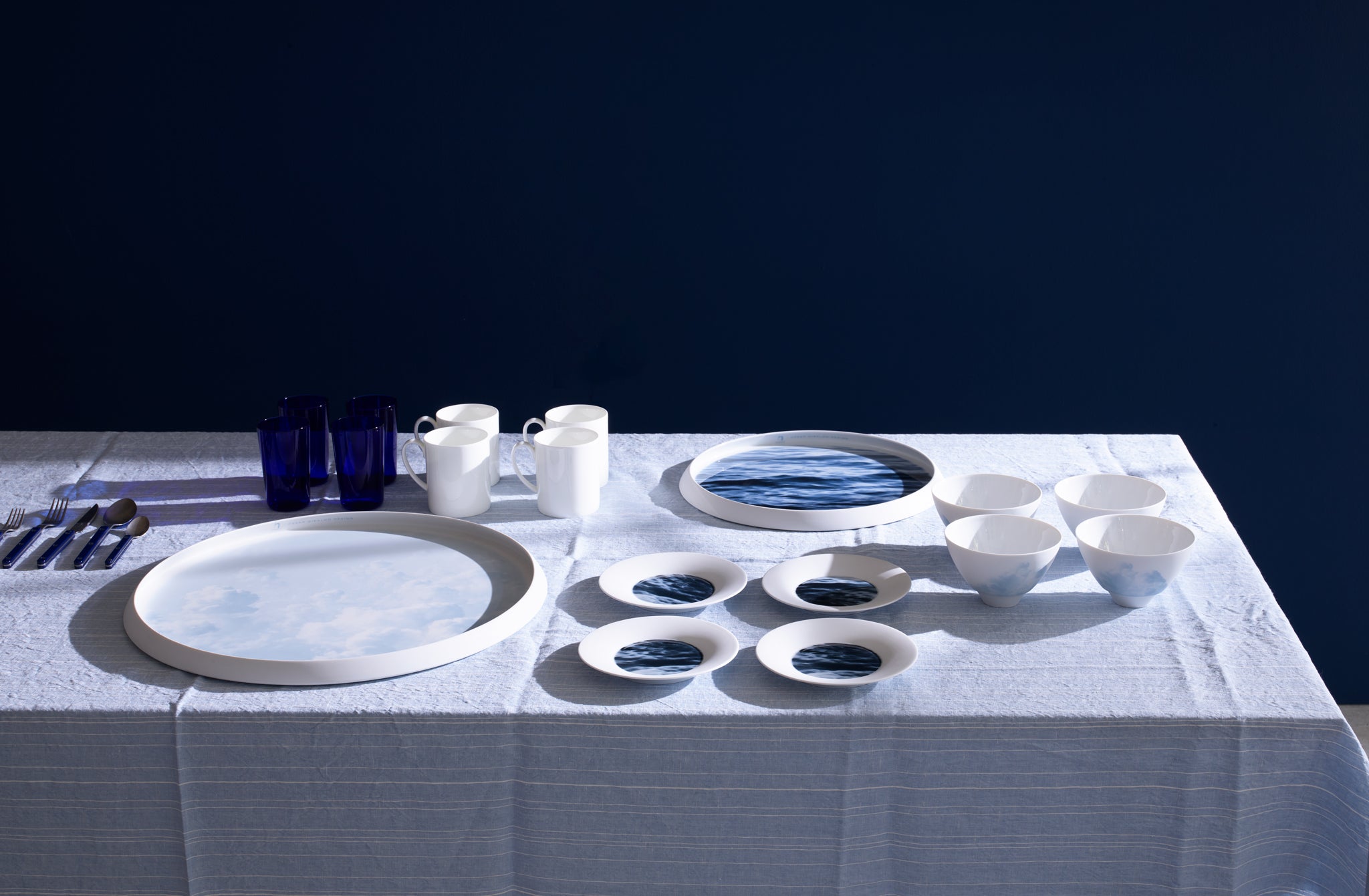 Photo realistic blue and white porcelain dinnerware and large serving trays of waves and clouds set in a gridded pattern with white porcelain coffee cups and cobalt glasses atop a light blue Greta striped linen tablecloth by Chiarastella Cattana.