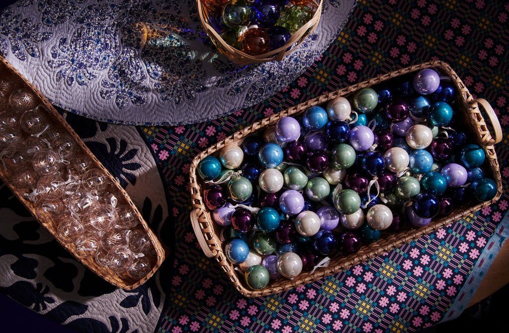 Glass holiday ornaments in baskets sitting on Hawaiian tree skirts and Gregory Parkinson night sky garden throw