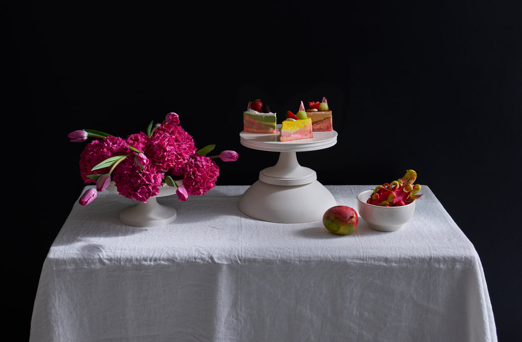 White John Julian serveware on a white table accented with vibrant pops of pink orange and yellow by the unique touch of mango dragon fruit marbelized cakes and a floral arrangement offset by a sophisticated black backdrop.
