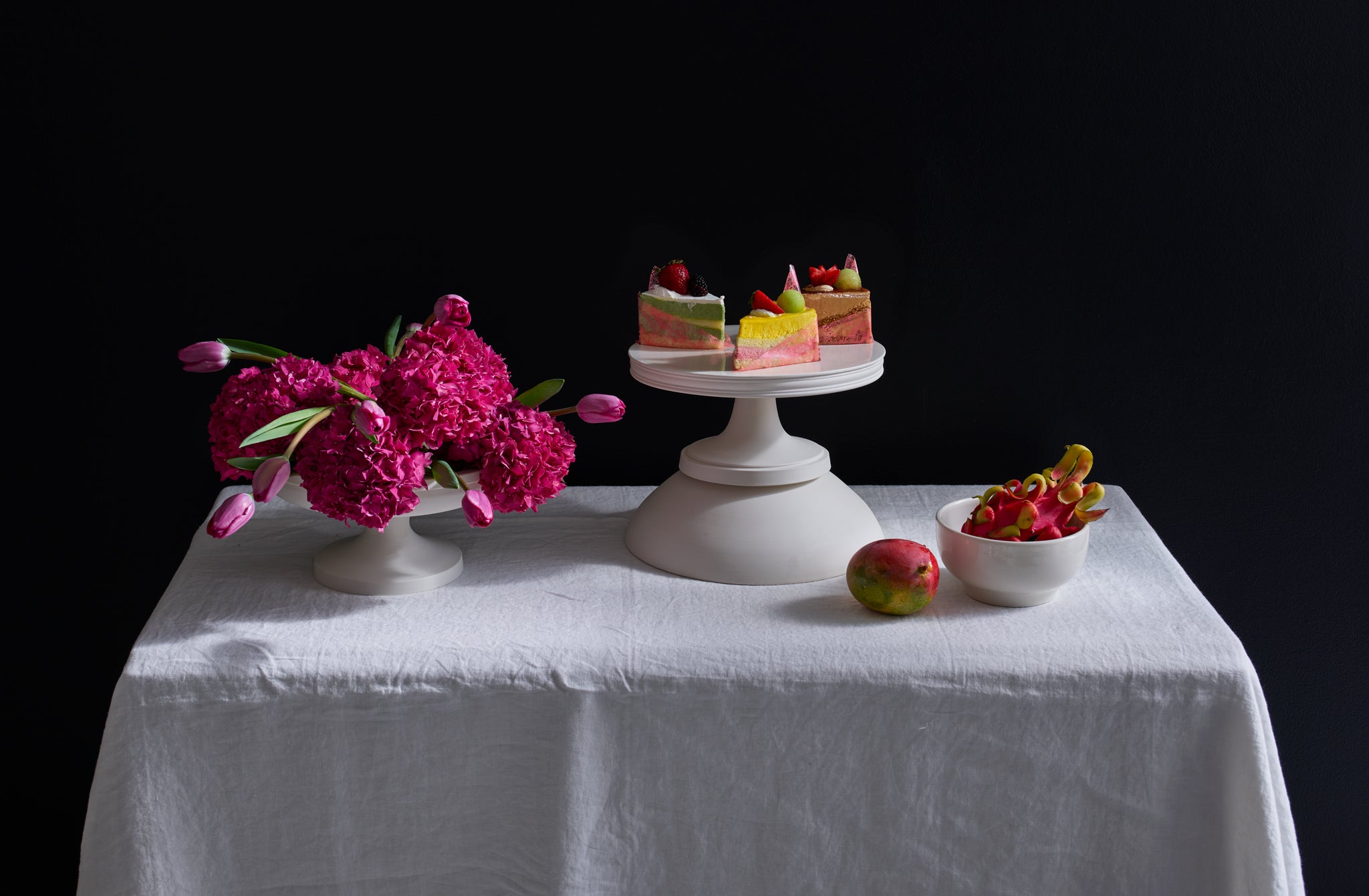 White John Julian serveware on a white table accented with vibrant pops of pink orange and yellow by the unique touch of mango dragon fruit marbelized cakes and a floral arrangement offset by a sophisticated black backdrop. Default