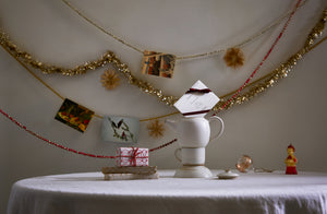 Sparkling garlands strung with straw star ornaments and seasonal greeting cards behind a round table with a playful stacks of Christiane Perrochon ceramics and small gifts.