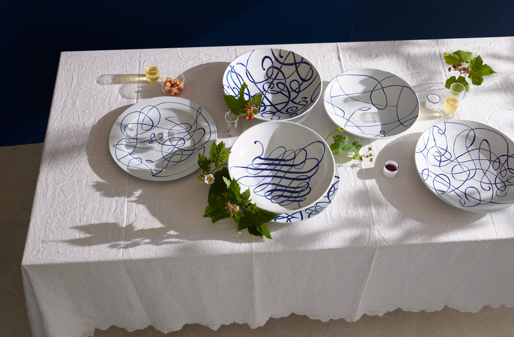 White linen tabletop seen from above and set with white hand-painted cobalt script ceramic platters and bowls by the artist Melissa Goldstein set with summer florals around and underneath.