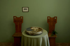 Two ninty sixties French carved chairs against a moss painted wall flank Michaël Verheyden marble Komm bowl sitting on table draped with moss macrame edge linen tablecloth.