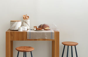 MARCH butcher block parson's table by Union Studio shown with Sawyer Made blue painted leg kitchen counter stool, and topped by linen kitchen towel and Michaël Verheyden Calcutta marble vessels holding utensils, and a breakfast of toast and honey.