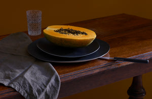 Stacked matte black Molosco dinnerware holding half of a bright papaya on an antique table with a Lobmeyr painted MARCH tumbler and charcoal heavy linen napkin. . Default