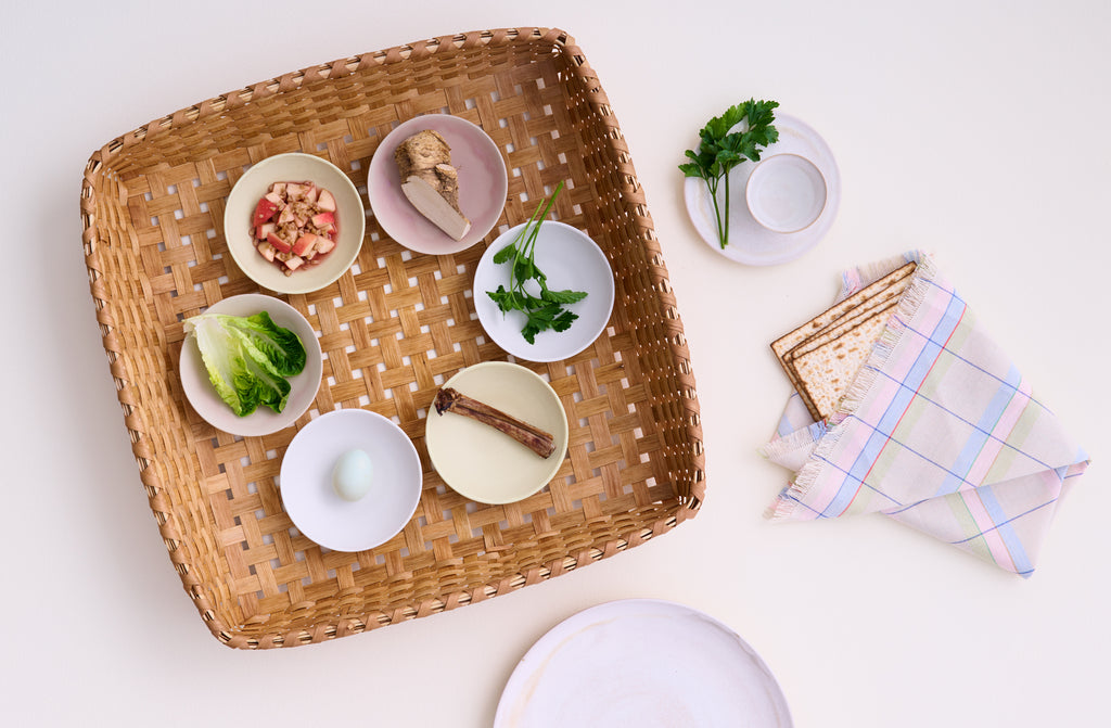 Default. Hand woven square natural black ash basket set with Christiane Perrochon spice bowls in dove, pale yellow and pink, as a Seder plate for passover with a pastel plaid napkin wrapping matzah bread.