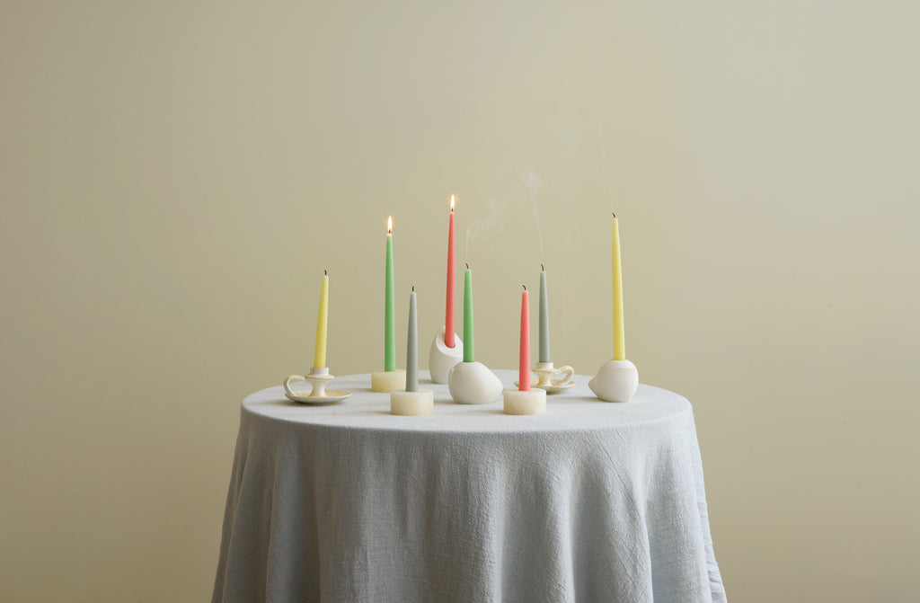 Round table covered with pale blue linen tablecloth and set with white and cream candle holders in onyx, splatterware and white gesso and filled with lit taper candles in pastel colors.