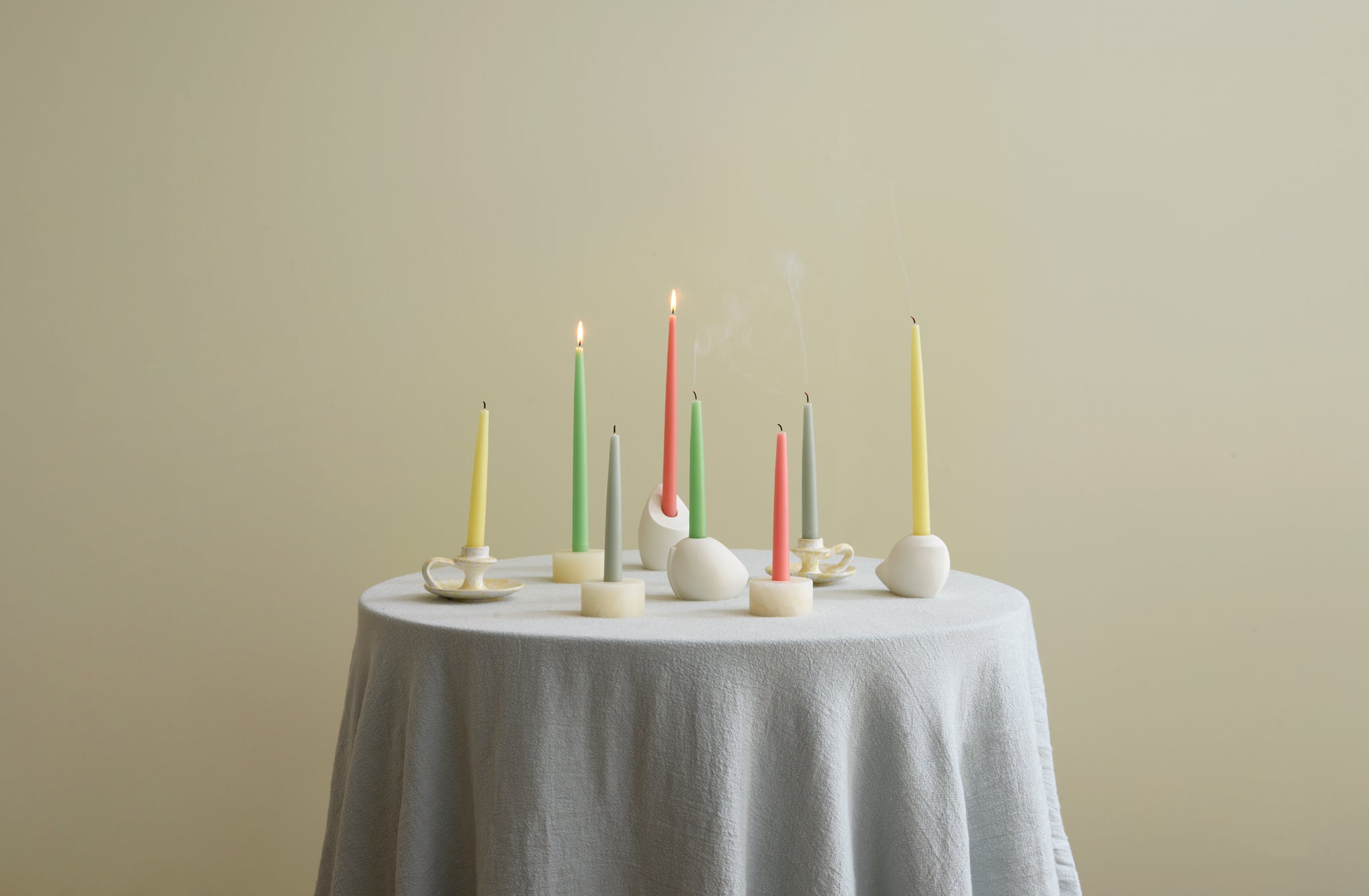 Round table covered with pale blue linen tablecloth and set with white and cream candle holders in onyx, splatterware and white gesso and filled with lit taper candles in pastel colors.