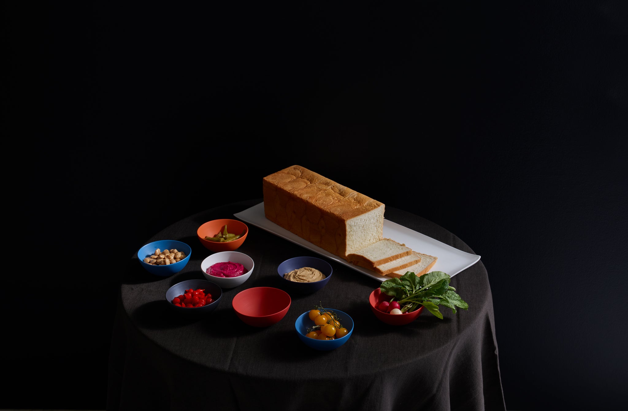 Table set like a stage with colorful Rina Menardi bowls filled with jewel-toned dips and snacks next to a white Rina Menardi tray holding a sculptural sliced loaf of bread.