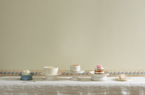 Cake stands by Christiane Perrochon, John Julian, and Ann Demeulemeester set with white cakes on a ivory linen windowpane tablecloth, antique pastel wallpaper border lines the pale yellow wall behind.