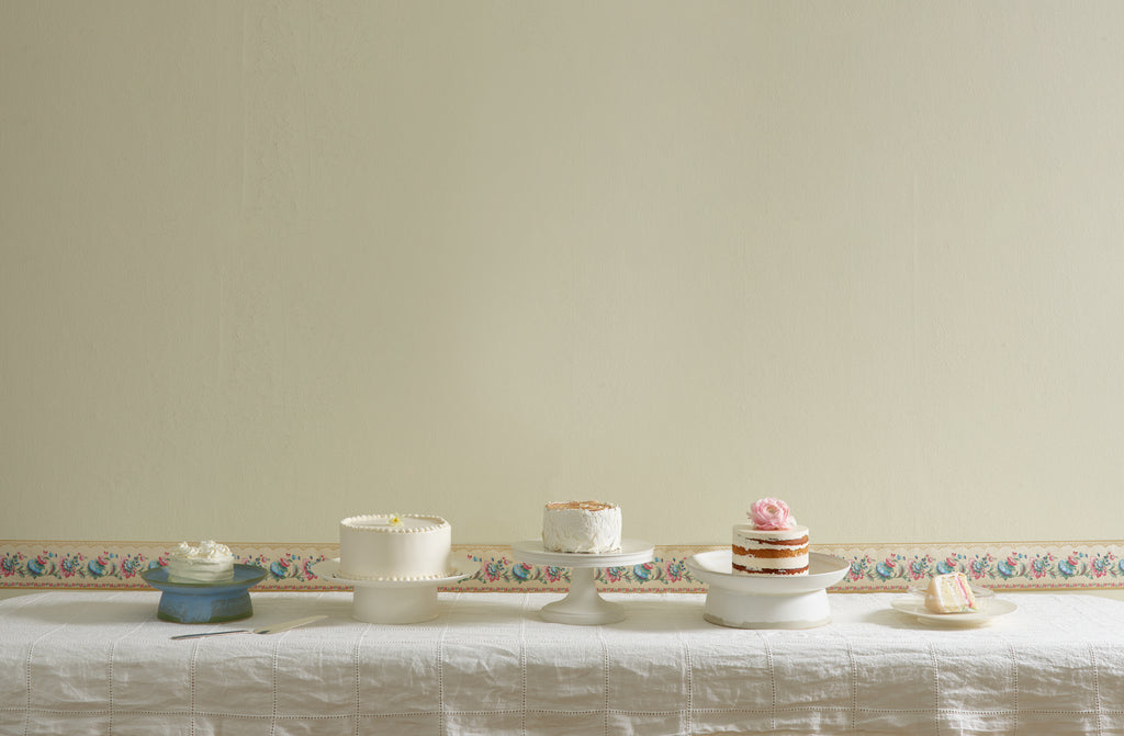 Cake stands by Christiane Perrochon, John Julian, and Ann Demeulemeester set with white cakes on a ivory linen windowpane tablecloth, antique pastel wallpaper border lines the pale yellow wall behind.