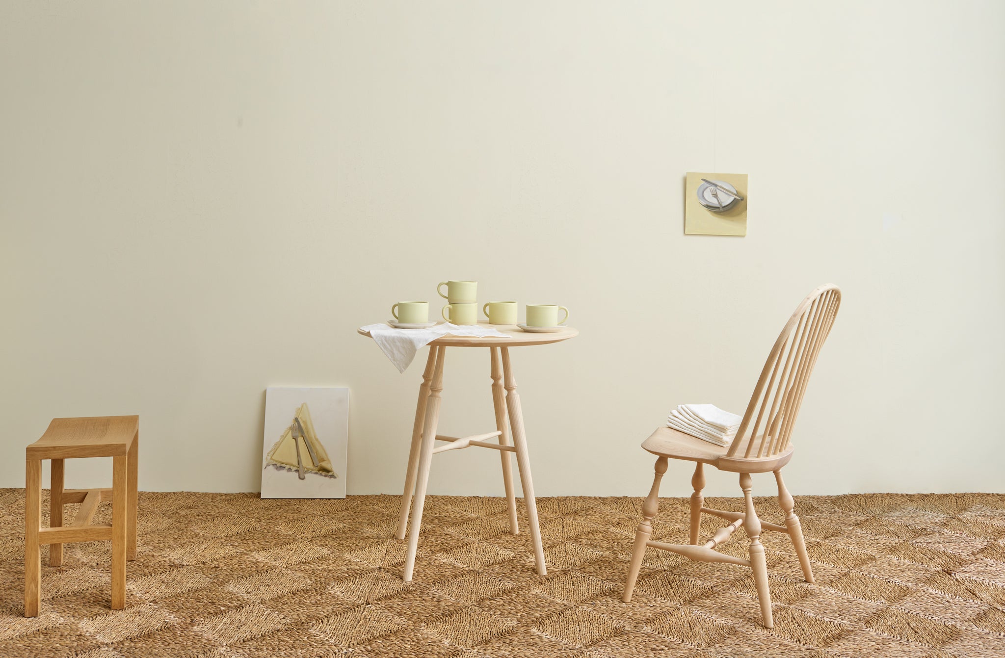 Room with rush woven rug set with Sawyer Made ash round table and windsor chair stacked with Christiane Perrochon pale yellow mugs, Carrie Mae Smith still life paintings seen in the background.