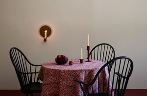 Sawyer made black windsor arm chairs set at round table covered with gregory parkinson red printed tablecloth and set with david chipperfield candlesticks and small bordeaux marble bowl with david chipperfield lit mirror sconce on the wall