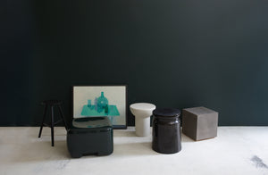 A selection of side tables that can be stools and stools that can be side tables in different materials and shapes from Faye Toogood Michael Verheyden Henry Dean and Sawyer Made next to a painting of green objects on a table by Jason Line.