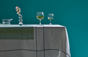 Grided linens layered on a table complemented by Yoshihiko Takahashi wine glasses and lipped bowl reflecting the light set against a teal backdrop.