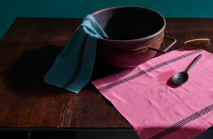 Turquoise and pink piano kitchen towels paired with an antique brass basin bowl highlighted in the light and an ebonized wood spoon resting close by. Default