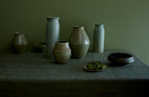 Victoria Morris and Christiane Perrochone ceramic vessels next to Brickett Davda plate holding green figs on homespun sage green tablecloth against a sage green wall.