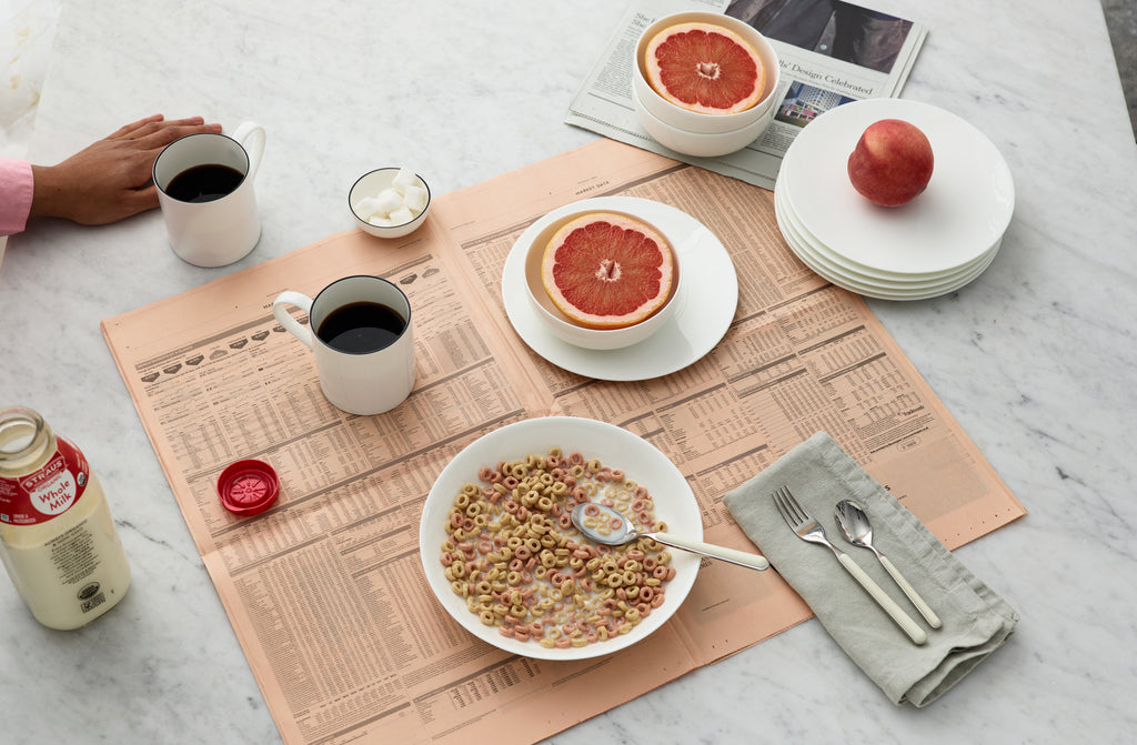 Marble kitchen counter set for breakfast with Dibbern white bone china plates and bowls filled with colorful cereal and cut pink grapefruit atop a financial times newspaper with Fantasia china white flatware on a pale green linen napkin. Default