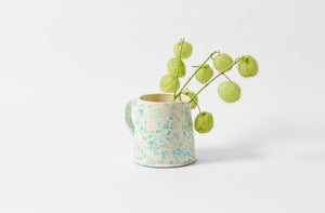 Turquoise and cream large splatterware pitcher holding bright green plant pods.