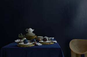christiane perrochon dinnerware and petite boule vases stacked atop a table covered in a navy linen tablecloth with rush placemats next to a faye toogood roly poly dining chair