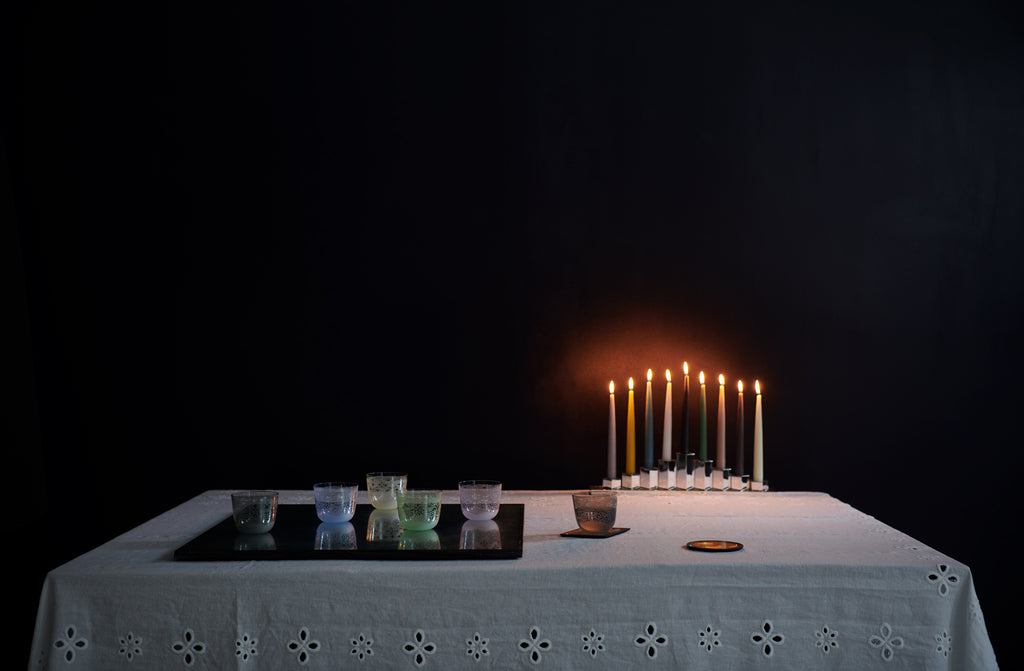 lobmeyr viennese lace engraved tumblers in various colors set on a mirrored surface with a lit candlelobra shown behind with multicolored tapered candles